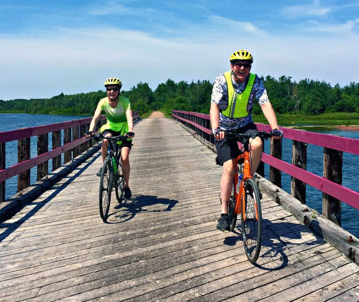 2 happy cyclists traversing a bridge along the traffic-free Confederation Trail, and bike paths of the National Park with our Confederation Trail Bike