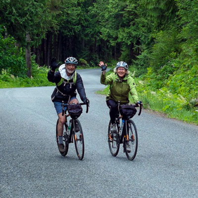 This image shows two individuals enjoying a scenic bicycle tour on a paved road surrounded by dense trees on Vancouver Island