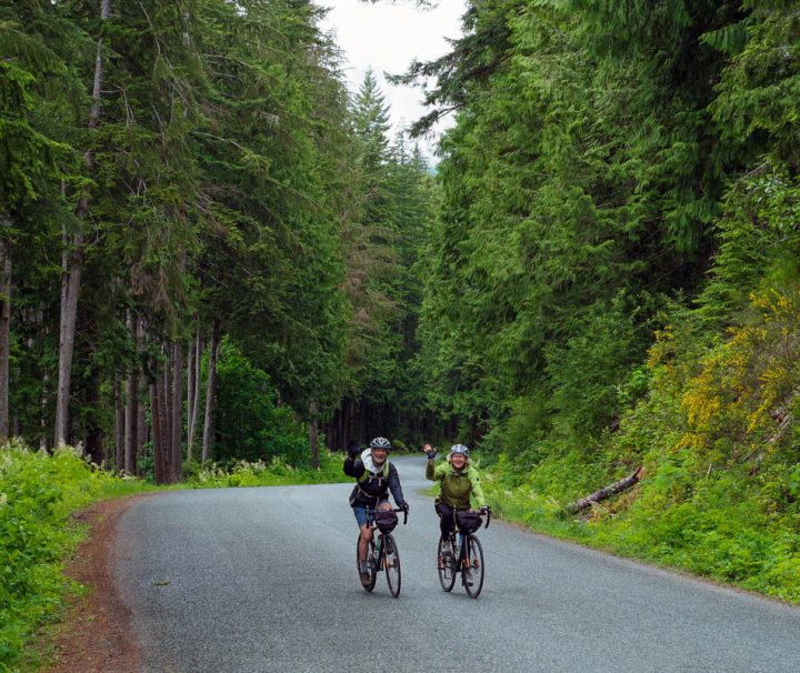 This image shows two individuals enjoying a scenic bicycle tour on a paved road surrounded by dense trees on Vancouver Island