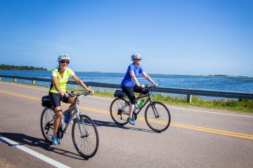 This image shows two individuals enjoying a scenic bicycle ride along a paved coastal side road of Prince Edward Island