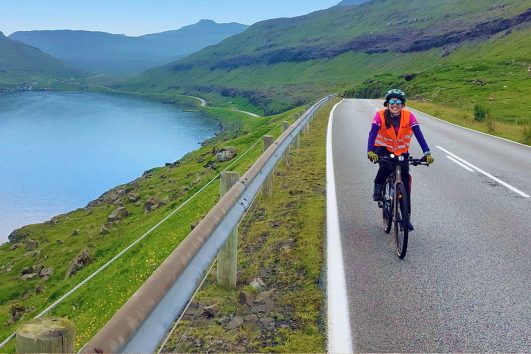 This image captures a moment of someone cycling on a scenic coastal road, in a mountainous region in the Faroe Islands