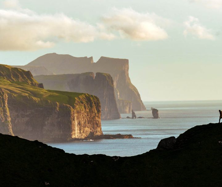 The image shows two people hiking on a mountainside overlooking dramatic cliffs and the ocean of the coast of Faroe Islands